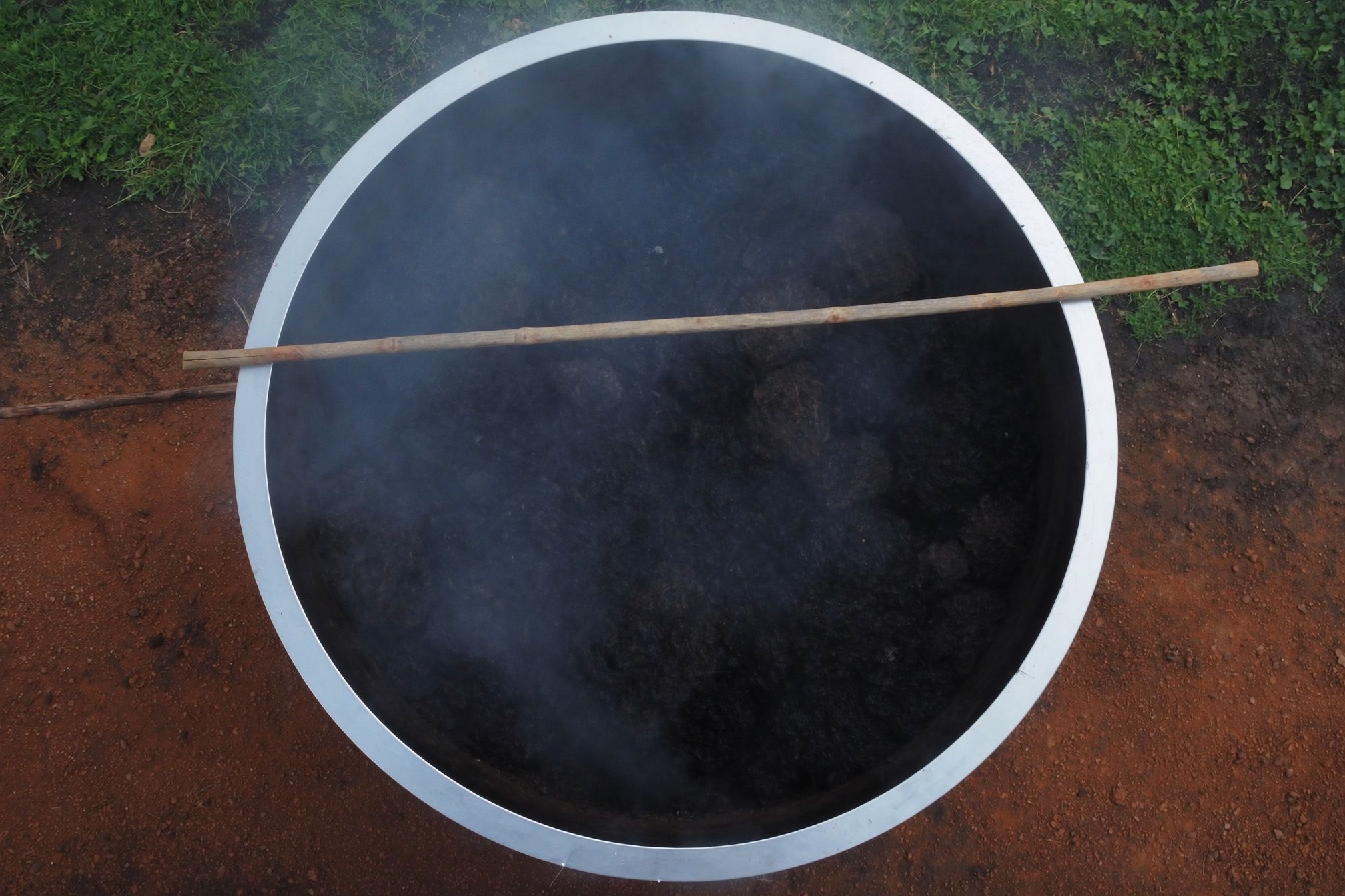 Looking down into a wide steel-rimmed vessel of dark converting biomass with steam lifting off it and a wooden paddle resting across the rim.