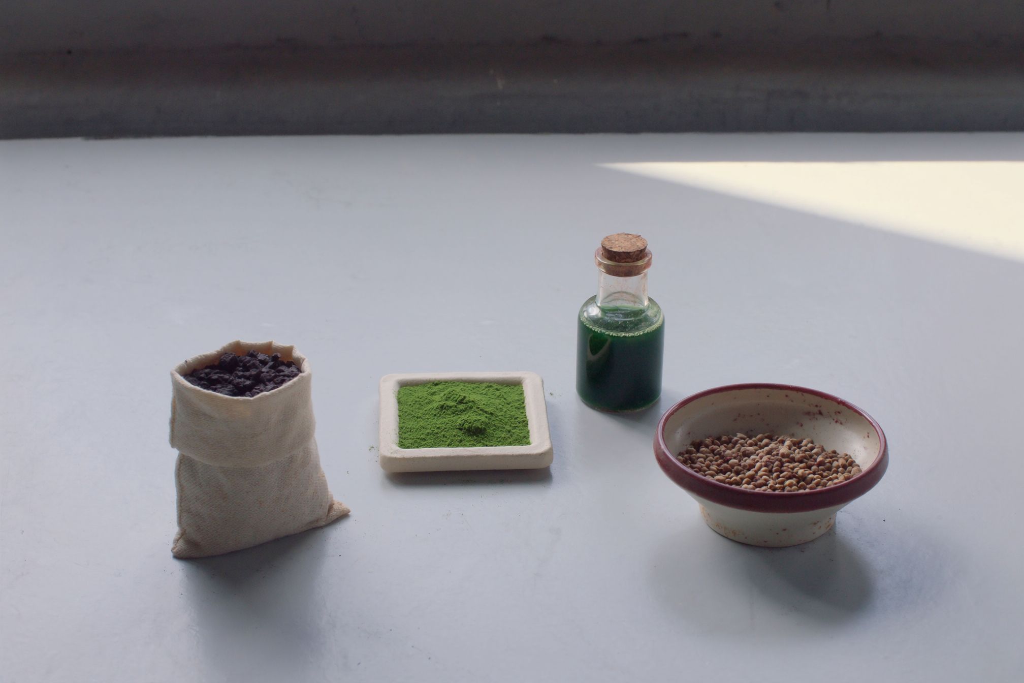 A plain pale surface holding an open sack of dark granular material, a shallow dish of bright green powder, a corked bottle of green extract and a bowl of feed pellets.
